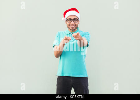 Hey you! Its christmas man! Bearded man in santa hat and glasses, pointing finger and looking at camera with toothy smile. On gray background. Indoor, Stock Photo