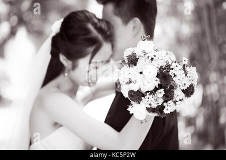 young asian newly wed couple hugging each other during wedding ceremony, focus on the bouquet, black and white. Stock Photo