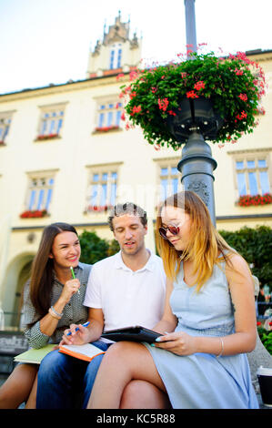 Three students are preparing for exams. Two girls and a guy sitting on a bench and talking animatedly. In the hands of their notes and their tablet. Stock Photo