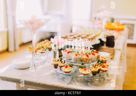 Delicious wedding cupcakes are laying on the dessert stand while the pink marshmallows in the huge vase. The wedding table set Stock Photo