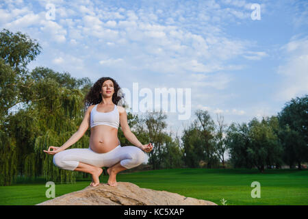 Pregnant woman in meditation pose on stone grass in the park. Green lawn and sky in the background. Stock Photo