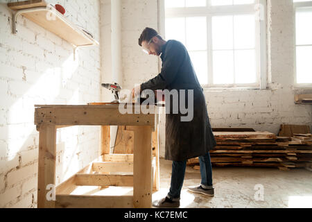 Joiner using electric tools while working in carpentry. Millennial worker getting on-the-job trailing in woodworking workshop, improve his skills in p Stock Photo