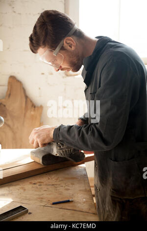Skilled professional carpenter in workwear and protective eyewear sanding wooden plank, using electric belt sander, custom woodworking job, young craf Stock Photo