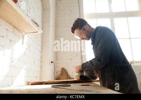 Carpenter in protective glasses polishes wooden board with handle belt sanding machine on workbench in workshop. Small business owner working in his m Stock Photo