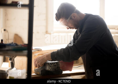 Side view of young joiner wearing protective glasses for safety sanding planks, skilled workman carpenter woodworking in small workshop with equipment Stock Photo