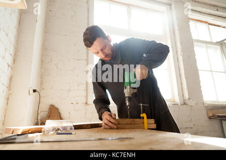 Skilled professional carpenter in workwear using electric drill machine, custom woodworking, making woodware, young experienced craftsman drilling woo Stock Photo
