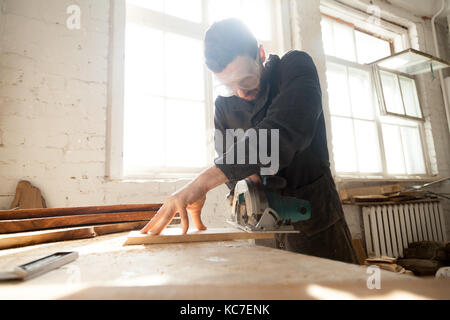 Carpenter cutting wooden board on workbench with handle circular saw. Entrepreneur working on his small woodworking venture. Woodworker works on local Stock Photo