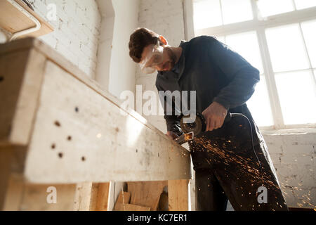 Worker in protective eyewear working in small workshop interior using die angle grinder for cutting steel bar, throwing sparks from metal grinding mac Stock Photo