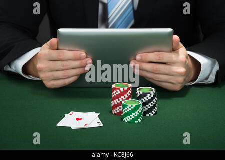 Poker Hand Holding Digital Tablet With Chips And Cards On Table Stock Photo