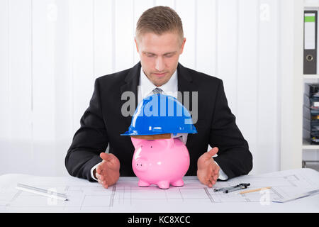 Young Male Architect With Piggybank Wearing Construction Helmet On Blueprint At Desk In Office Stock Photo