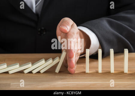 Close-up Of Young Businessperson Stopping Dominos From Falling On Wooden Desk Stock Photo