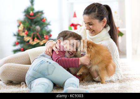 Happy child and his mother are lying on floor near Christmas tree and embracing dog. They are looking at pet and smiling. Stock Photo