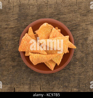 Top view of a small red clay bowl filled with cheese flavored cone shaped corn chips on an old brown board. Stock Photo