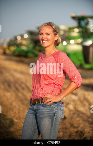 A YOUNG FEMALE FARMER STANDS IN A FIELD NEAR JOHN DEERE COMBINES PRIOR TO HARVESTING WHEAT ON THE FAMILY FARM NEAR BRECKENRIDGE, NORTH DAKOTA Stock Photo