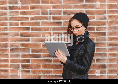 Waist portrait of young woman standing in the street beside brick wall, listening music online with headphones from tablet. Happy beautiful woman usin Stock Photo
