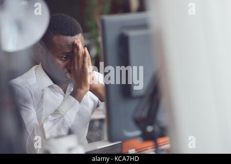 Tired African man sitting at a office after a hard workday, working on laptop, trying to concentrate Stock Photo