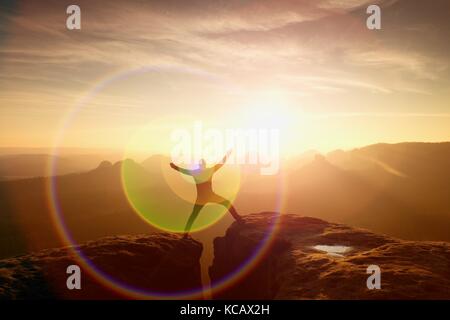 Strong light reflection in lens. Jumping hiker in black celebrate triumph between two rocky peaks. Wonderful daybreak. Stock Photo