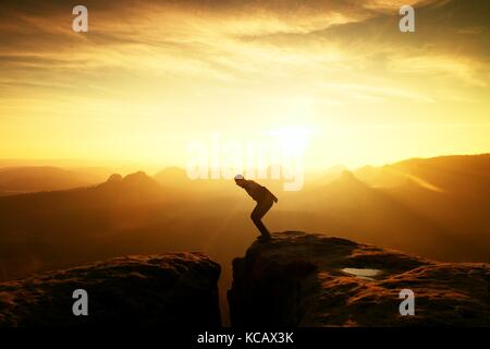 Man in black jumping between rocky empires. Dreamy daybreak in sandstone mountains, heavy orange mist in deep valley. Stock Photo