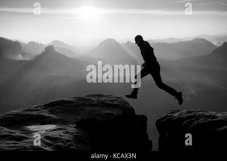 Hiker in black is jumping between rocky peaks. Amazing activities in rocky mountains, heavy mist in deep valley. Black and white photo Stock Photo