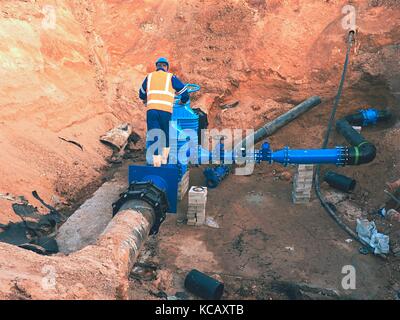 Worker in safety clothes drive valve conduit  on City potable water pipe  joined with black eccentric fastener members into old pipeline system. Stock Photo