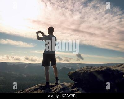 Man in tshirt and shorts takes photos with smart phone on peak of rock empire. Dreamy fogy landscape, spring orange pink misty sunrise in a beautiful  Stock Photo