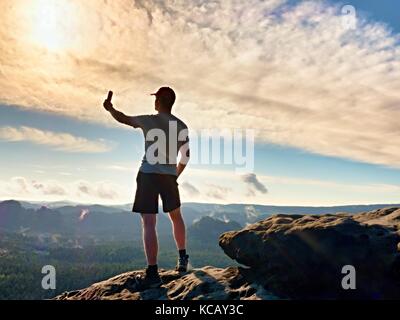 Man in tshirt and shorts takes photos with smart phone on peak of rock empire. Dreamy fogy landscape, spring orange pink misty sunrise in a beautiful  Stock Photo