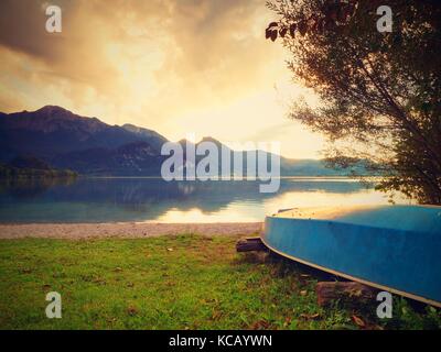 Upside down fishing paddle boat on bank of Alps lake. Smooth levelof autumnal lake. Dramatic and picturesque scene. Mountains in water mirror. Stock Photo