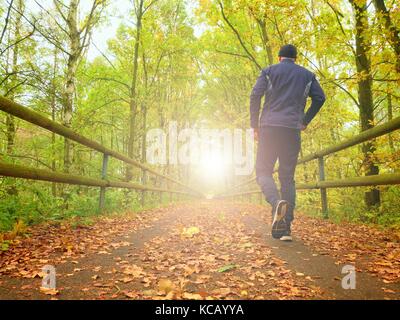 Flare, reflection. Sportsman in  black sortswear run on road. The man is slowly running on asphalt way covered by autumn leaves. Pathway in park, beec Stock Photo