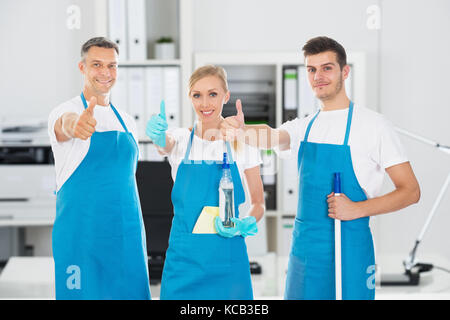 Portrait Of Smiling Janitors Holding Cleaning Equipments Showing Thumbs Up In Office Stock Photo