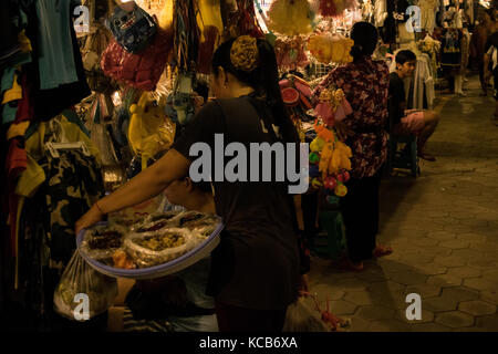 Asian woman selling Asian finger food and treats at Phnom Penh night market, talking to a woman at her clothes stall, next to a toys stall. Cambodia Stock Photo