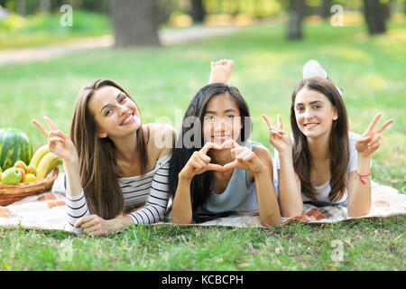 Portrait of three young women showing sign peace and heart. One Asian girl and two Caucasian having fun outdoors. Female friendship concept. Models sm Stock Photo