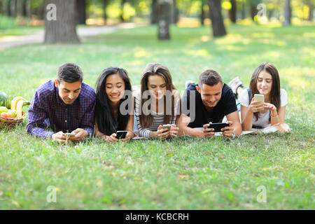 Five young friends lying on grass and using mobile phones. Two men and three women resting outdoors and browsing in smartphones, exchanging photos, ch Stock Photo