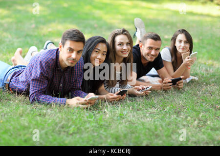 Five young friends lying on grass and using mobile phones. Two men and three women resting outdoors and browsing in smartphones, exchanging photos, ch Stock Photo