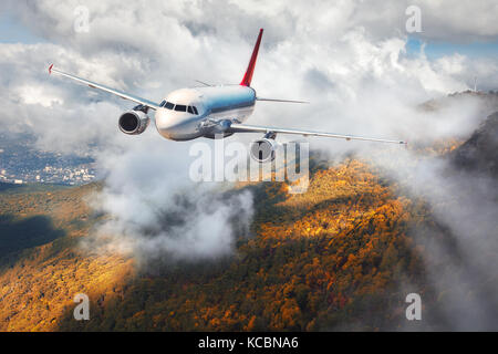 Airplane is flying in clouds over mountains with autumn forest at sunset. Landscape with passenger airplane, cloudy sky and trees. Passenger aircraft 