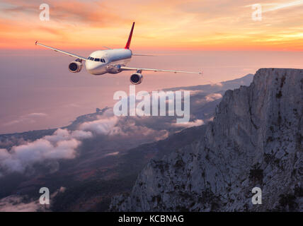Airplane is flying over rocks and clouds at sunset. Landscape with passenger airplane, mountains, sea and orange cloudy sky in summer. Passenger aircr Stock Photo