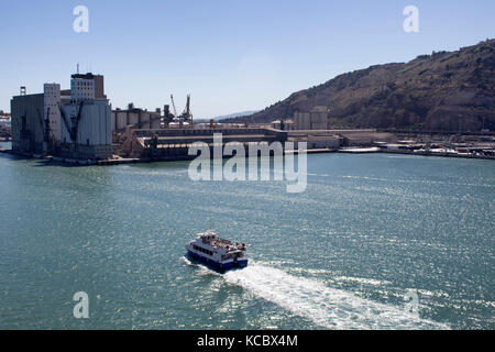 Aerial view of a tour boat with people leaving Barcelona port. Shipping / logistic facilities are in the background. It's great commercial importance  Stock Photo