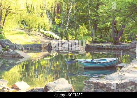 Sunny spring day in the city park with a pond surrounded by rocks. Trees reflection in the water. Blue wooden boat with two paddles peacefully resting Stock Photo