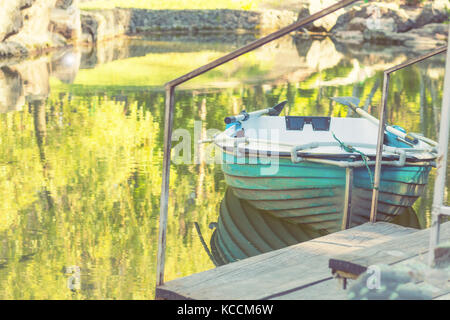 Sunny spring day in the city park with a pond surrounded by rocks. Trees reflection in the water. Blue wooden boat with two paddles peacefully resting Stock Photo