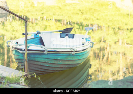 Sunny spring day in the city park with a pond surrounded by rocks. Trees reflection in the water. Blue wooden boat with two paddles peacefully resting Stock Photo