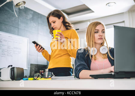 Two woman in the office, one is using laptop and other standing and using smart phone and holding cup of coffee Stock Photo