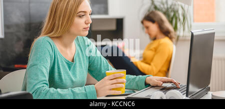 Young business woman sitting at the desk, drinking coffee and using laptop in the background is a woman by the desk taking notes Stock Photo