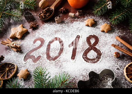 New Year 2018 written on flour and Christmas Decorations Gingerbread cookies, cinnamin, oranges, spices, nuts and cookie cutters on wooden background. Stock Photo