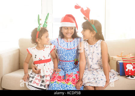 Happy Indian family celebrating Christmas holidays, with gift box and santa hat sitting on couch at home, adorable Asian children on festival mood. Stock Photo