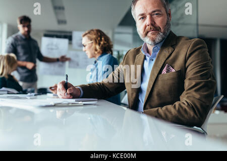 Mature businessman sitting in conference room with colleagues in background. Senior man sitting in board room during presentation. Stock Photo