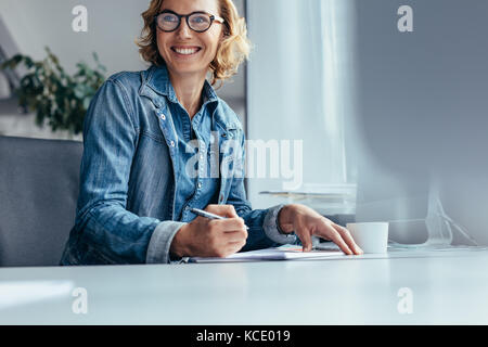 Beautiful young businesswoman sitting in office looking away and smiling. Female executive at work. Stock Photo