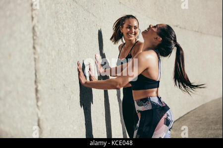 Two young woman doing stretching workout against a wall and smiling. Female friends exercising together in morning outdoors. Stock Photo