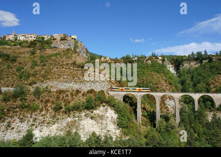 Picturesque landscape with a commuter train on a stone viaduct below a perched medieval village. Méailles, Alpes-de-Haute-Provence, France.
