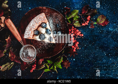 Close-up of a piece of pumpkin pie with powdered sugar and a strainer on a wooden plates. Handmade dishes in autumn flat lay. Conceptual stylised food still life, table top shot on dark background. Stock Photo