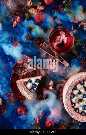 Dark autumn still life with a whipped cream blueberry cake, hot tea and floral decoration on a background with leaves and smoke. Conceptual stylized still life, table top shot on dark background. Stock Photo