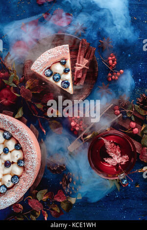 Dark autumn still life with a whipped cream blueberry cake, hot tea and floral decoration on a background with leaves and smoke. Conceptual stylized still life, table top shot on dark background. Stock Photo